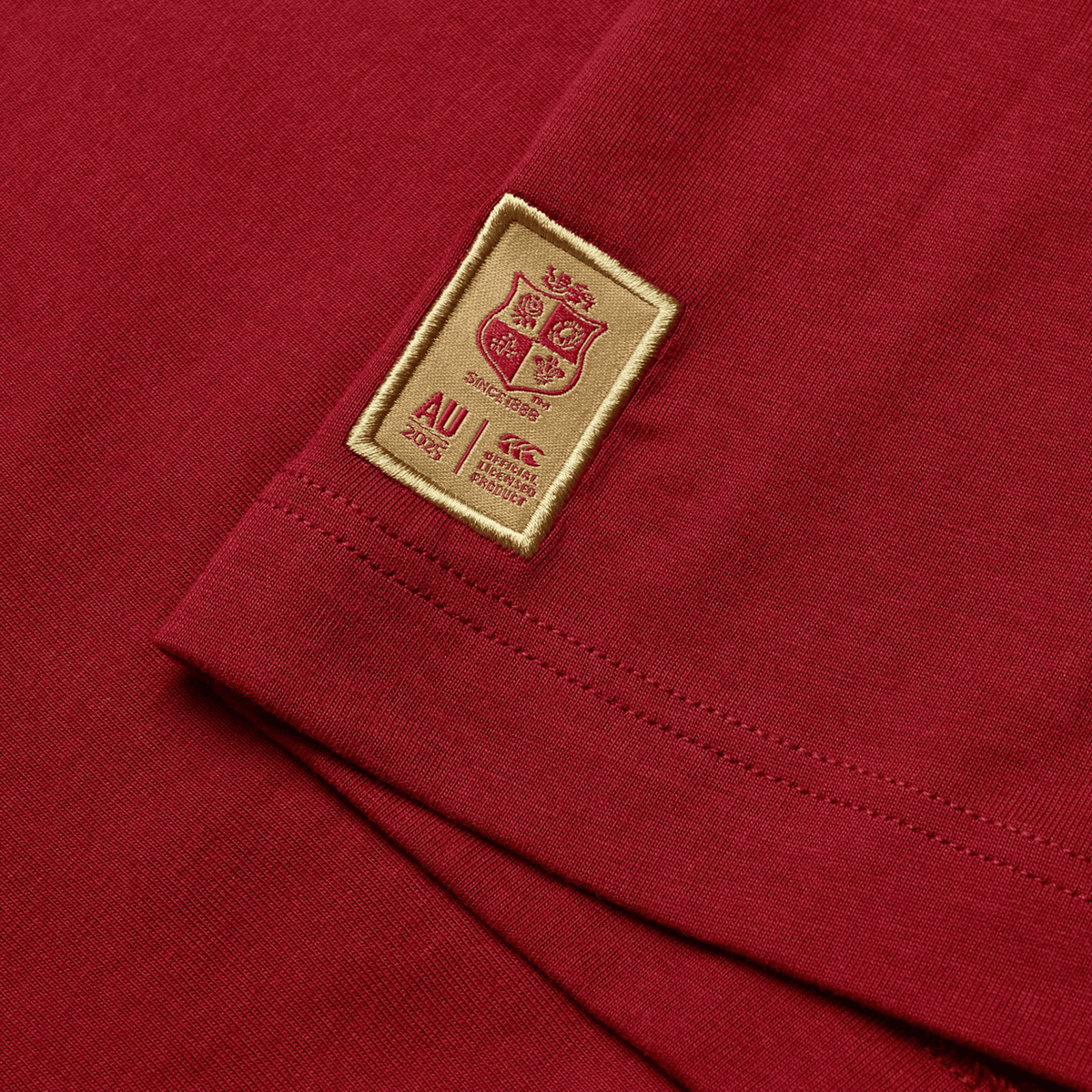 A close-up of the British &amp; Irish Lions 2025 Cotton Jersey Tee by Canterbury showcases a stitched patch on red fabric, featuring a coat of arms, the letters "AU," and the year "2025." Crafted from organic cotton, this tee is ideal for any rugby supporter, embodying the enduring spirit of the British &amp; Irish Lions.