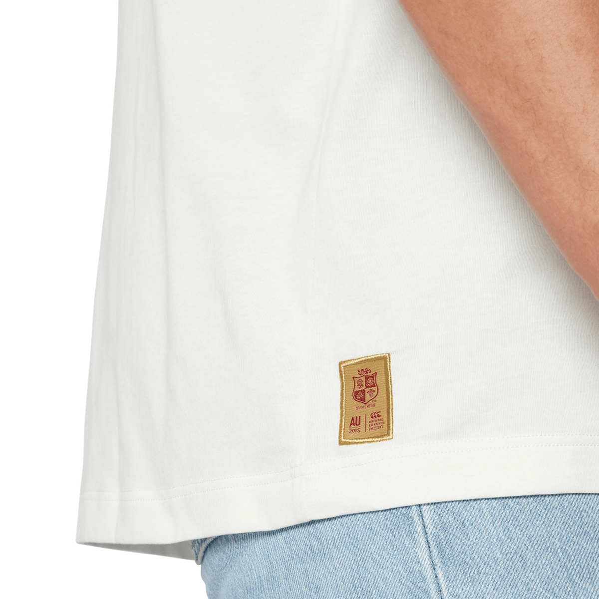 Close-up of a person wearing the British &amp; Irish Lions 2025 Cotton Jersey Tee by Canterbury in white, paired with light blue jeans. A small label featuring a red emblem and lettering is visible near the hem of the shirt, evoking a rugby supporter's pride.