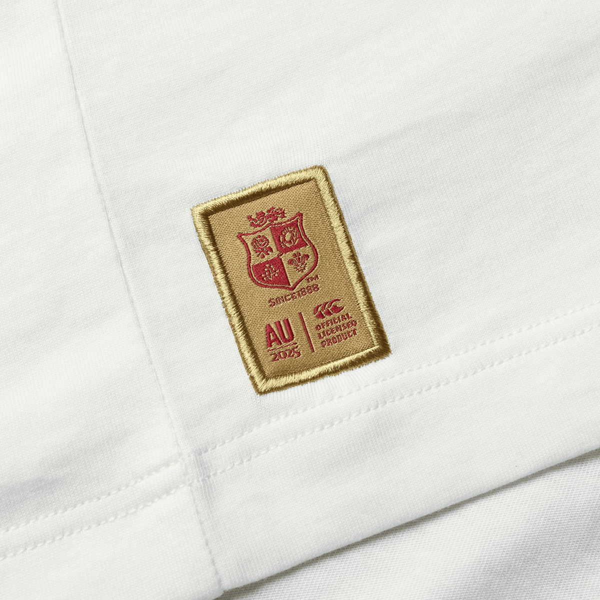 Made from organic cotton, this white fabric boasts a sewn patch with a red and gold emblem that reads "AU 2025" alongside additional details, signifying an official product endorsement. Ideal for any rugby fan wanting to express their support. This is the British &amp; Irish Lions 2025 Cotton Jersey Tee by Canterbury.
