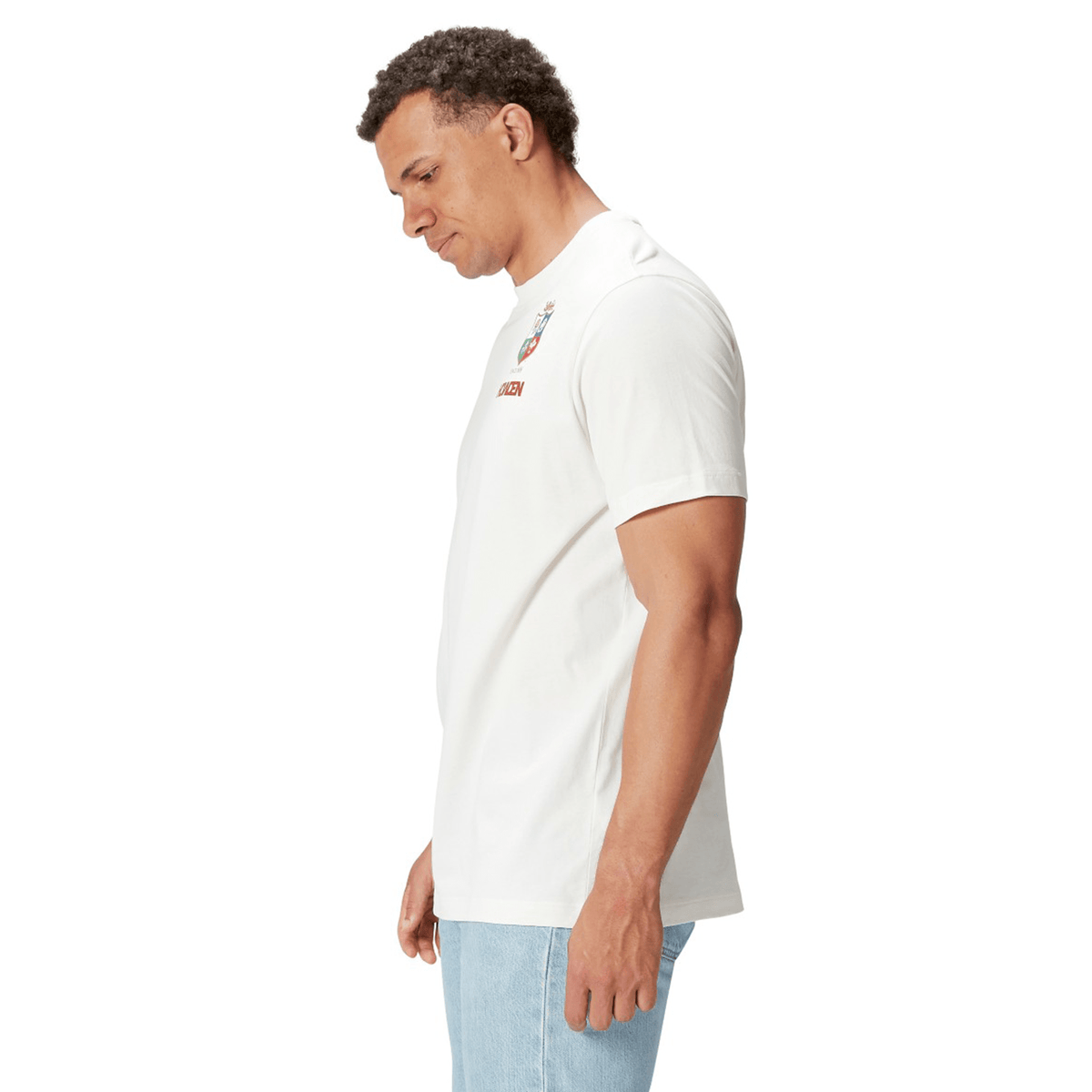 A rugby supporter in the British &amp; Irish Lions 2025 Cotton Jersey Tee by Canterbury, made from organic cotton, and blue jeans stands against a white background, facing left with a neutral expression.