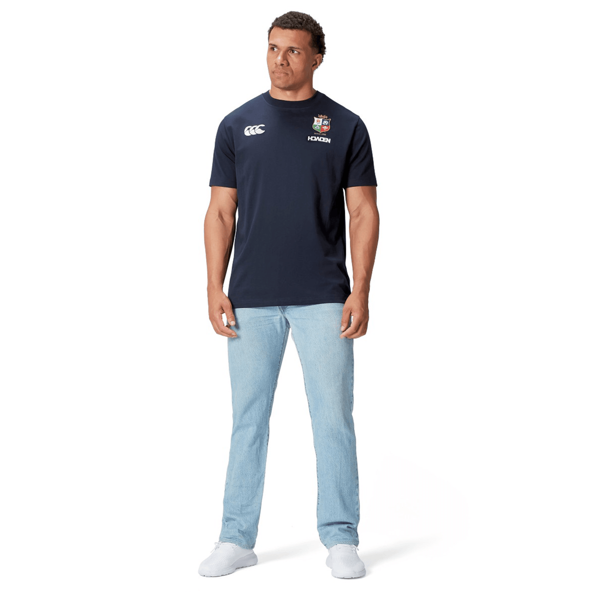 A rugby fan stands against a crisp white background, stylishly dressed in light blue jeans paired with the British &amp; Irish Lions 2025 Cotton Jersey Tee by Canterbury, featuring logos and crafted from organic cotton.
