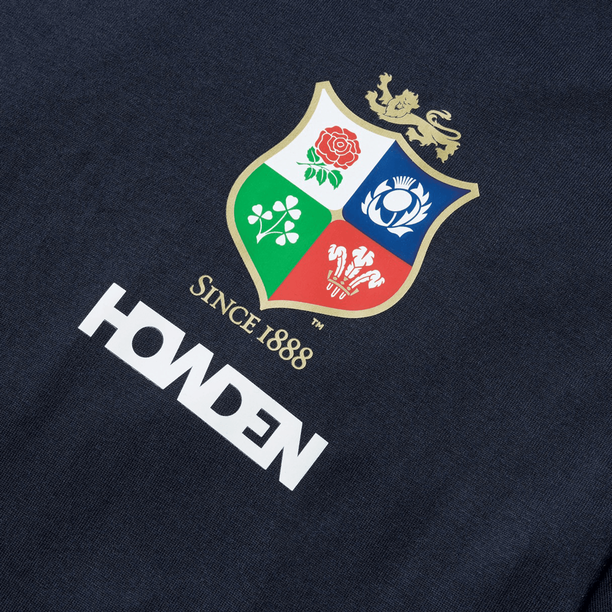 The British &amp; Irish Lions 2025 Cotton Jersey Tee by Canterbury features a shield emblem adorned with a rose, shamrock, thistle, and dragon, accompanied by the phrases "Since 1888" and "Howden" in bold white letters. Made from organic cotton, this navy shirt delivers comfort and style for rugby enthusiasts.