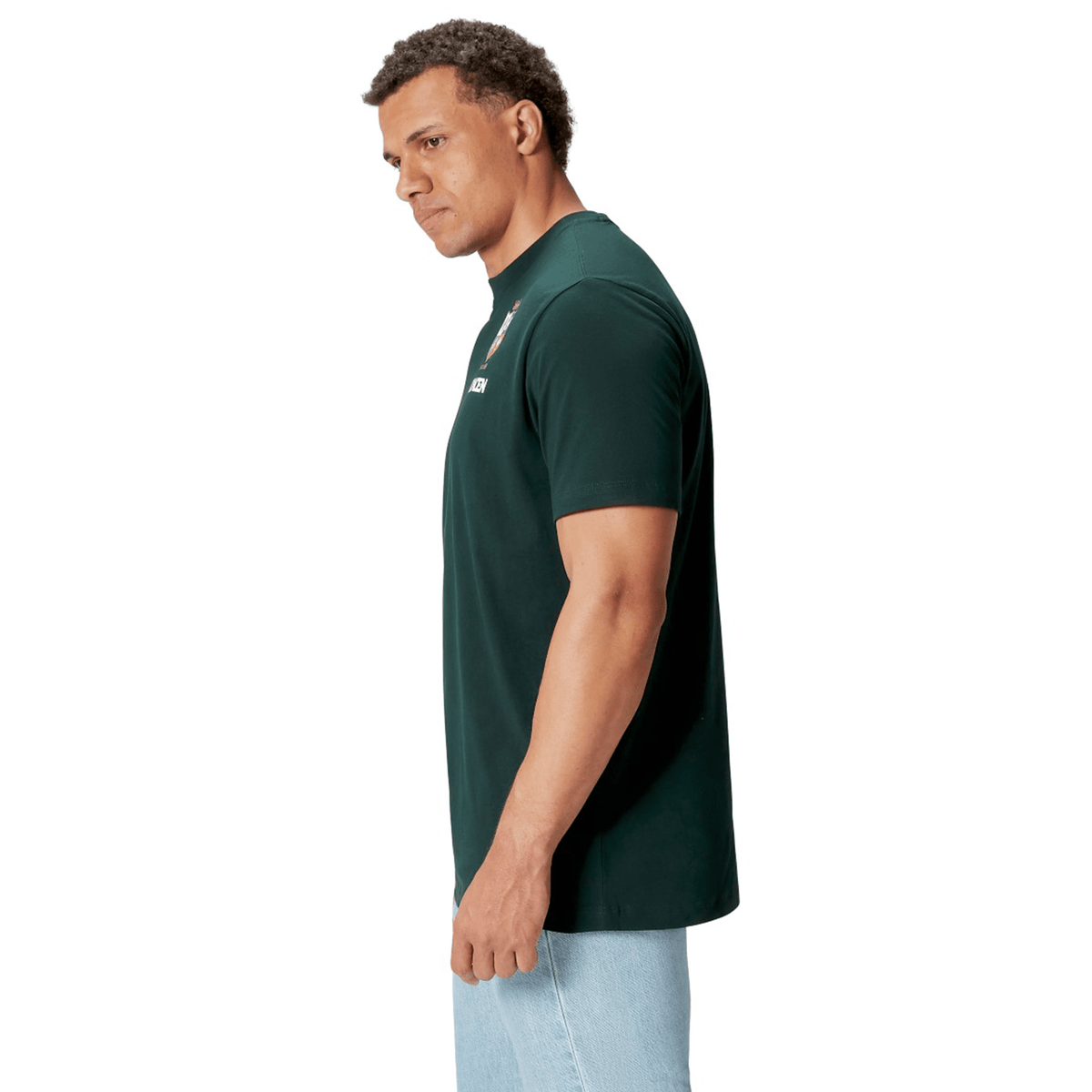 A rugby supporter wearing the British &amp; Irish Lions 2025 Cotton Jersey Tee by Canterbury in a dark green shade, paired with light blue jeans, is viewed from the side against a plain white background.