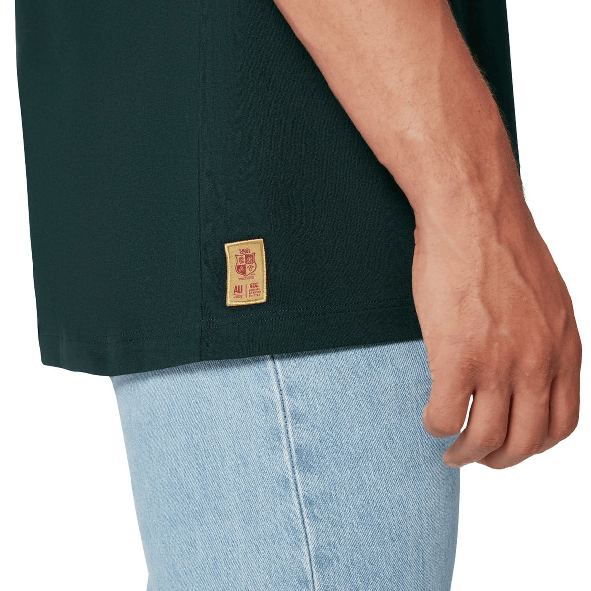 A person wearing the British &amp; Irish Lions 2025 Cotton Jersey Tee by Canterbury, crafted from organic cotton in dark green with a discreet tag, paired with light blue jeans. The focus is on the torso and right hand, exuding the casual yet spirited vibe of a rugby supporter.