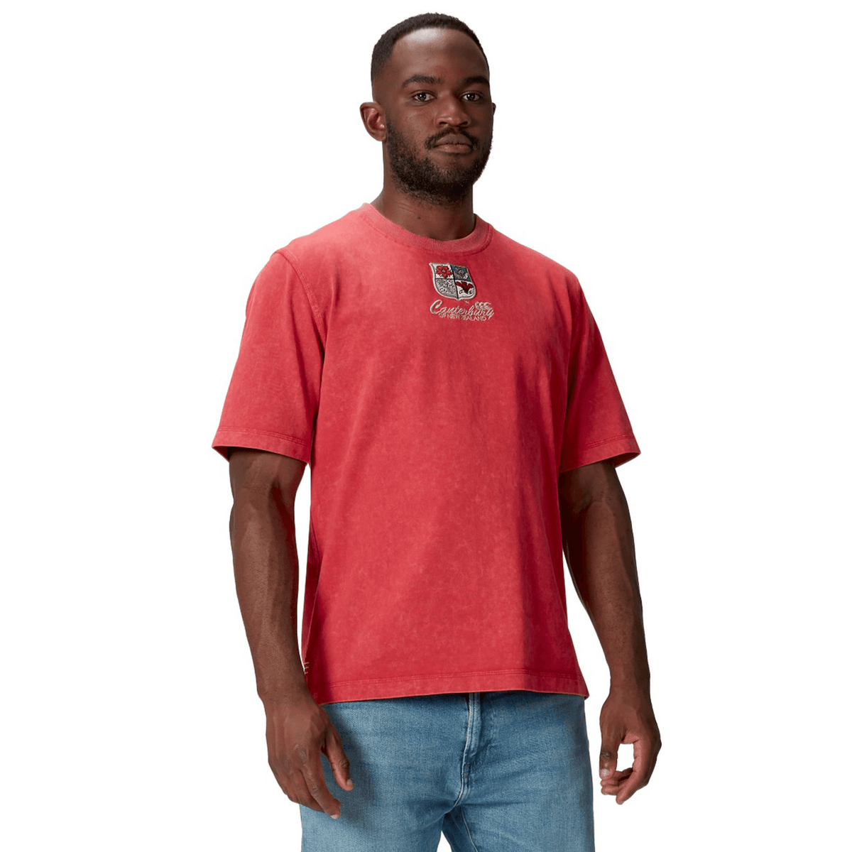 A man wears a British &amp; Irish Lions 25 Heritage Tee by Canterbury in red, matched with blue jeans, standing confidently against a plain white background.