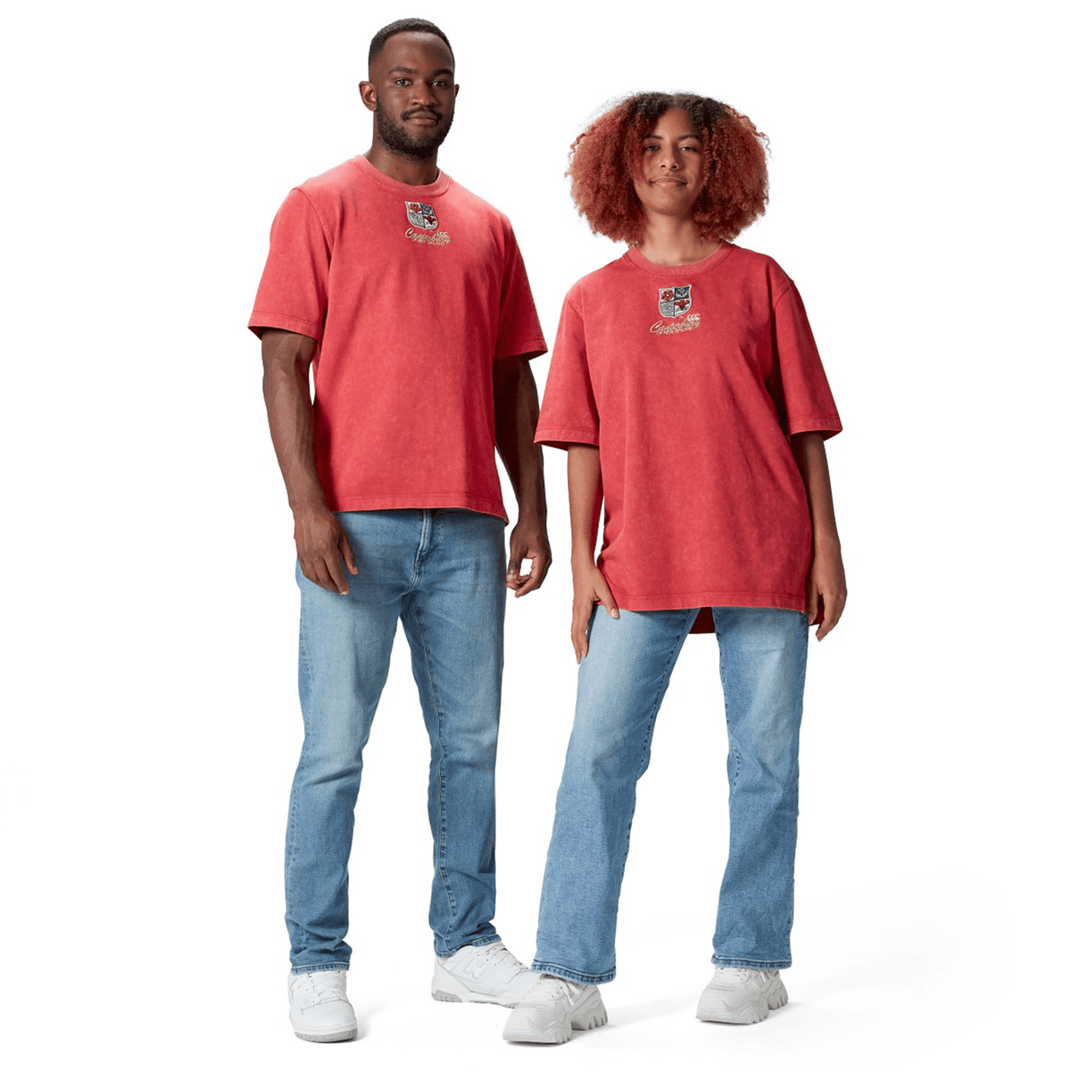 A man and a woman stand side by side, both dressed in red British &amp; Irish Lions 25 Heritage Tees by Canterbury, featuring a graphic design and made from soft organic cotton, complemented by light blue jeans.