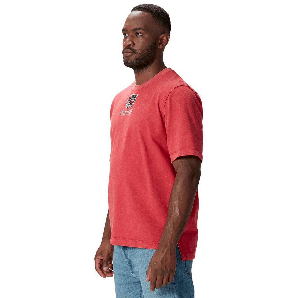 A man in a red British &amp; Irish Lions 25 Heritage Tee by Canterbury, showcasing a small graphic on the chest, paired with light blue jeans, stands against a plain white background.