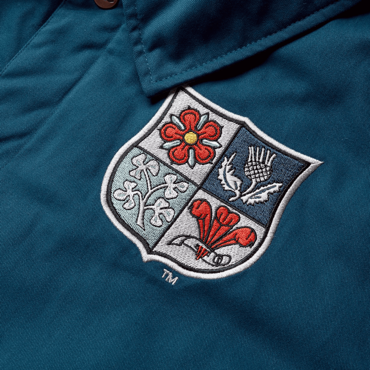 A close-up of the British &amp; Irish Lions 2025 Heritage Quilted Jacket by Canterbury in blue, featuring an embroidered shield logo with a red rose, thistle, shamrock, and daffodil—symbolic emblems of the British &amp; Irish Lions.