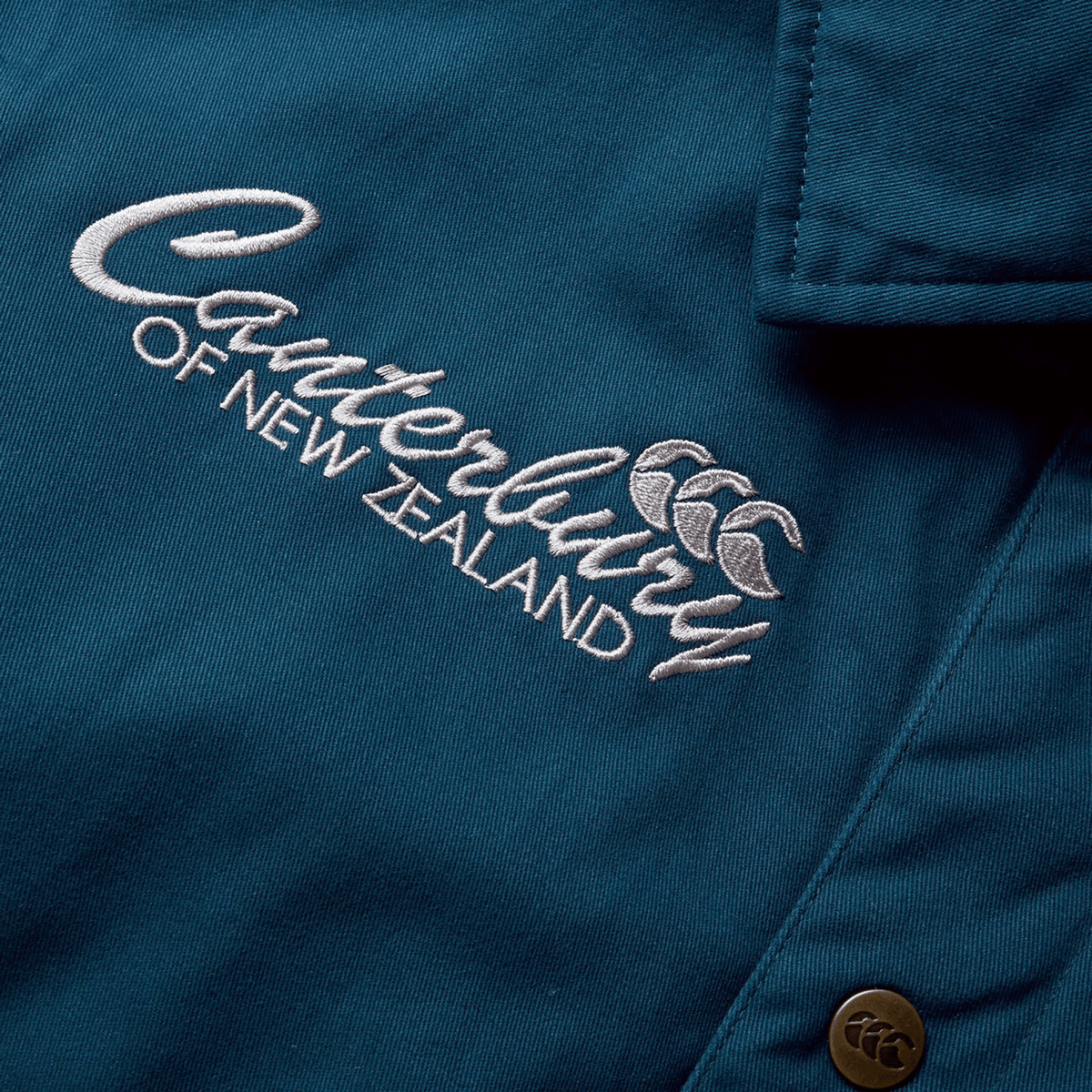A close-up of the Canterbury logo, embroidered in white on the blue fabric of the British &amp; Irish Lions 2025 Heritage Quilted Jacket by Canterbury, with a metal button visible—an iconic symbol celebrating the legacy of the Lions.