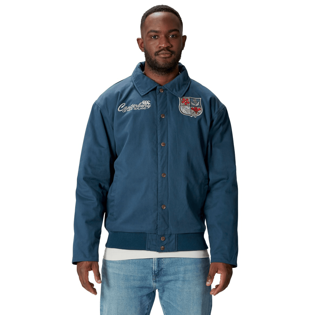 A man wearing a blue British &amp; Irish Lions 2025 Heritage Quilted Jacket by Canterbury, embellished with patches, stands against a white background, showcasing the timeless style synonymous with Canterbury and the British &amp; Irish Lions.