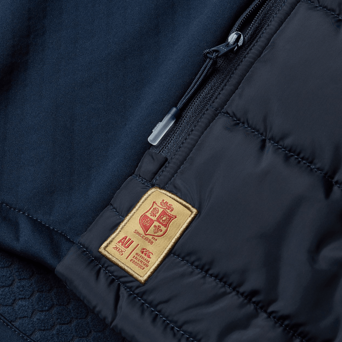 A close-up of the British &amp; Irish Lions 2025 Microlight Gilet by Canterbury showcases its black thermoreg fabric with a textured surface, accented by a small beige patch featuring red embroidered letters. The piece also includes a partially open zippered pocket, blending sleek design with the sustainable appeal of recycled polyester.