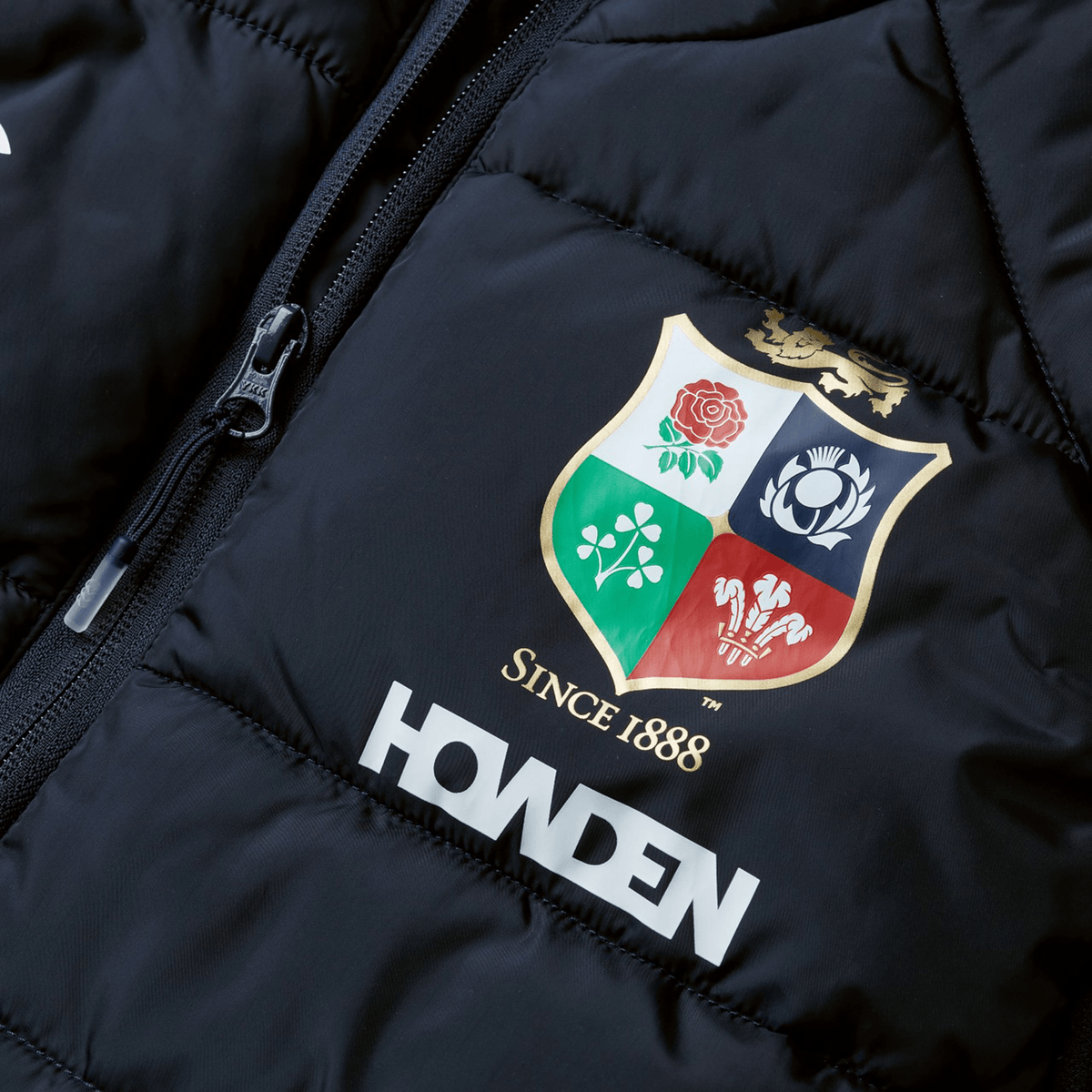 Close-up of a black British &amp; Irish Lions 2025 Microlight Gilet by Canterbury, crafted from recycled polyester. It showcases a colorful embroidered crest with floral motifs and the text "Since 1888," with the word "HOWDEN" printed below the crest.