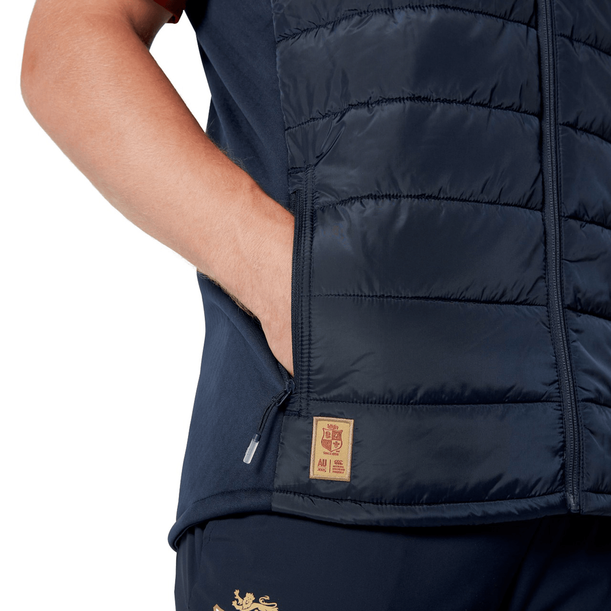 A person is wearing a navy blue British &amp; Irish Lions 2025 Microlight Gilet by Canterbury with matching pants, hands in pockets, and showcasing the logo patch. The ensemble is crafted from Recycled Polyester, offering both style and sustainability.