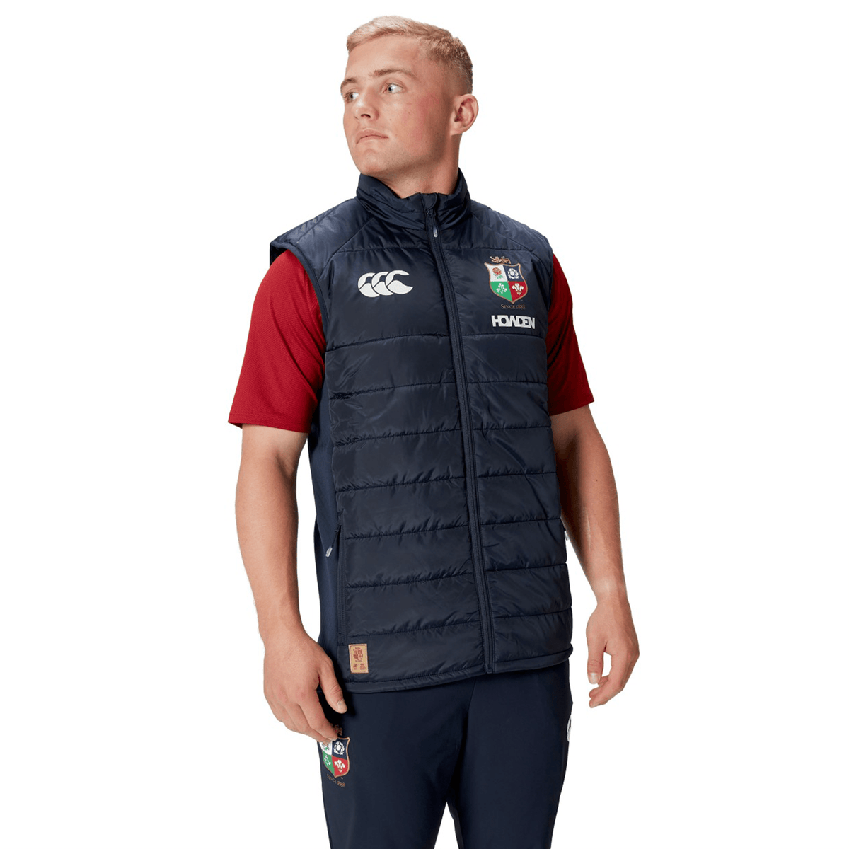 A person wearing a British &amp; Irish Lions 2025 Microlight Gilet by Canterbury in navy blue, made from recycled polyester, over a red shirt and navy pants, looks to the side.
