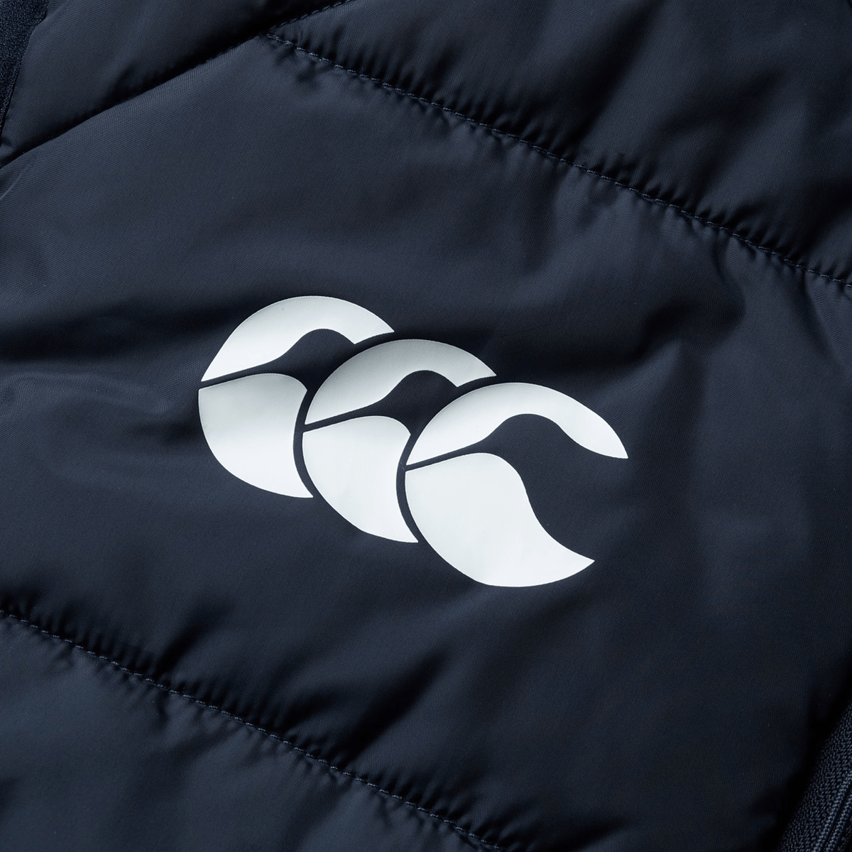 Close-up of the British &amp; Irish Lions 2025 Microlight Gilet by Canterbury, featuring a dark fabric with a white logo of three overlapping stylized wave shapes, made from recycled polyester.