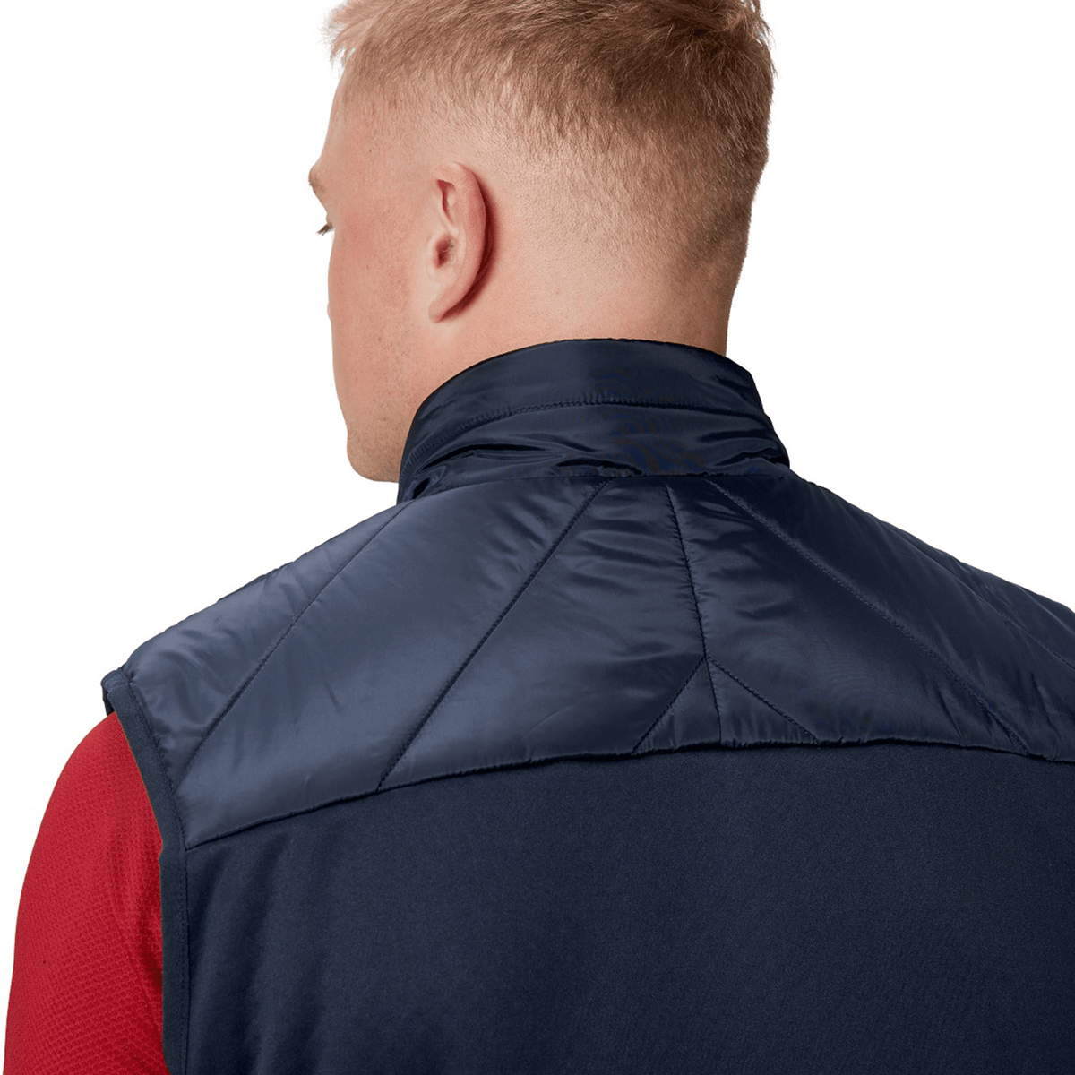 A person with short blonde hair is seen from behind wearing the British &amp; Irish Lions 2025 Microlight Gilet by Canterbury, crafted from recycled polyester in navy and black, over a red shirt.