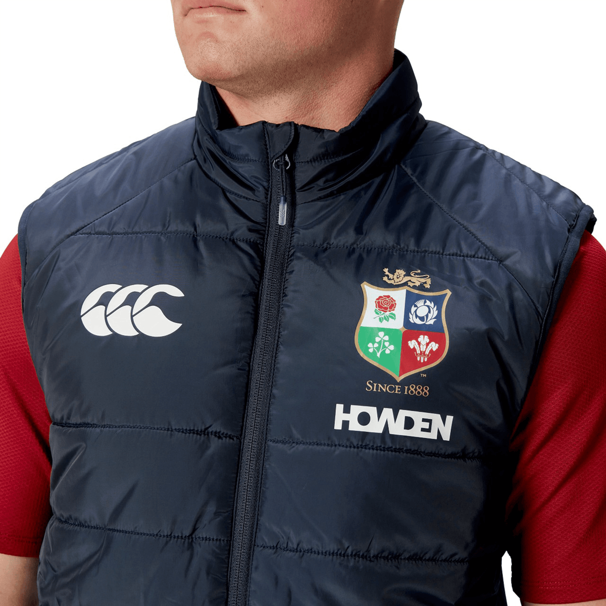 Dressed in a British &amp; Irish Lions 2025 Microlight Gilet by Canterbury, this individual showcases a navy blue vest adorned with the iconic Canterbury logo, the British and Irish Lions crest, and "HOWDEN" lettering. Made from recycled polyester with thermoreg technology, the gilet is fashionably paired over a red shirt.
