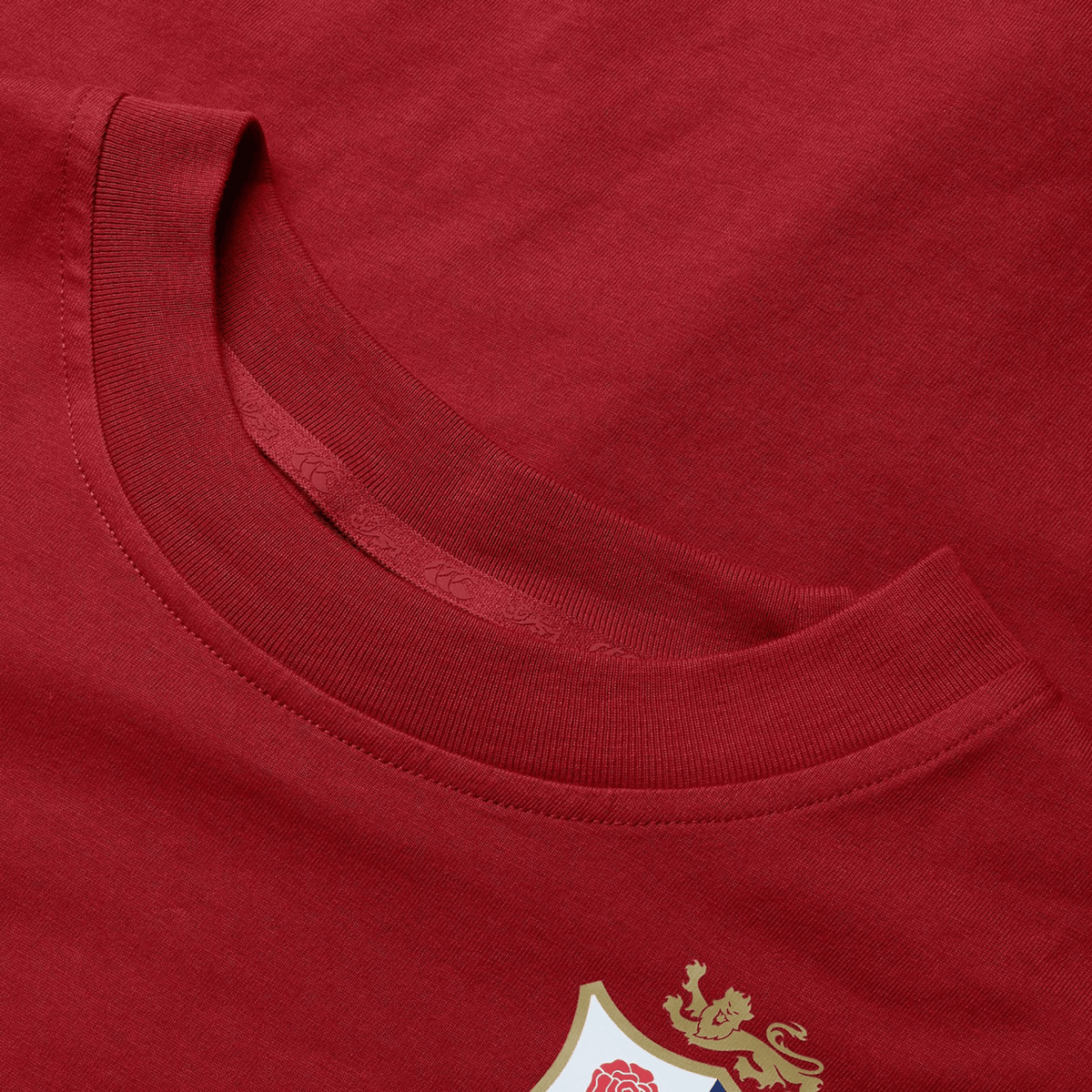 Close-up of the Women's British &amp; Irish Lions 25 Cotton Jersey Tee by Canterbury. This red T-shirt is crafted from organic cotton and features a distinctive shield emblem adorned with a white rose and gold lion in the lower corner, making it an ideal choice for any rugby supporter. The detailed design proudly highlights both the neckline and fabric texture.