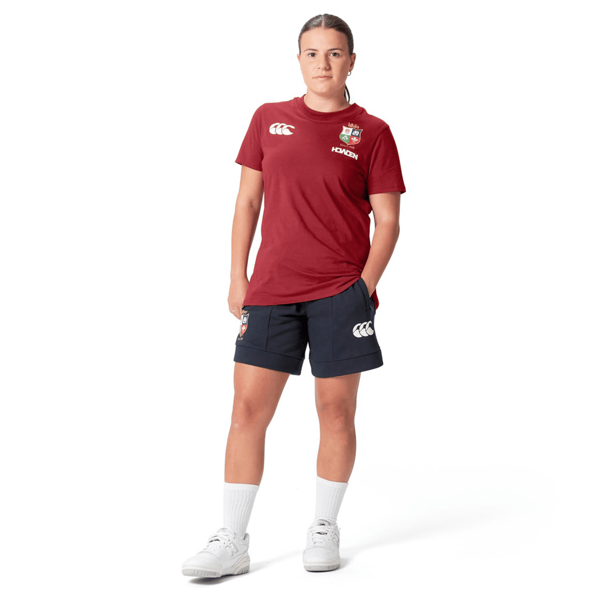 A person stands wearing a Women's British &amp; Irish Lions 25 Cotton Jersey Tee by Canterbury in red, along with navy shorts and white sneakers, against a white background.