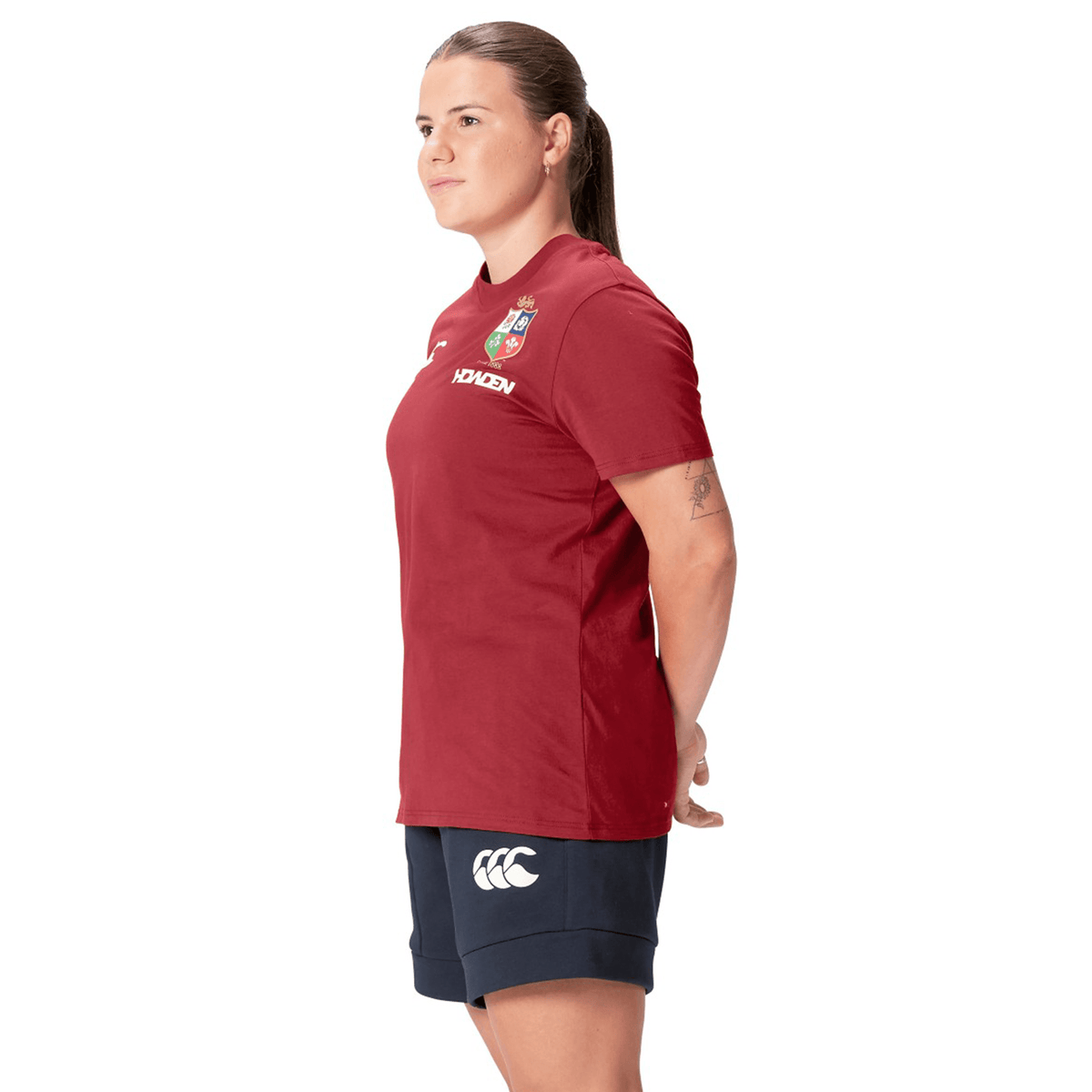 A rugby fan wearing the Women's British &amp; Irish Lions 25 Cotton Jersey Tee by Canterbury and navy shorts stands with their hands behind their back, facing left.