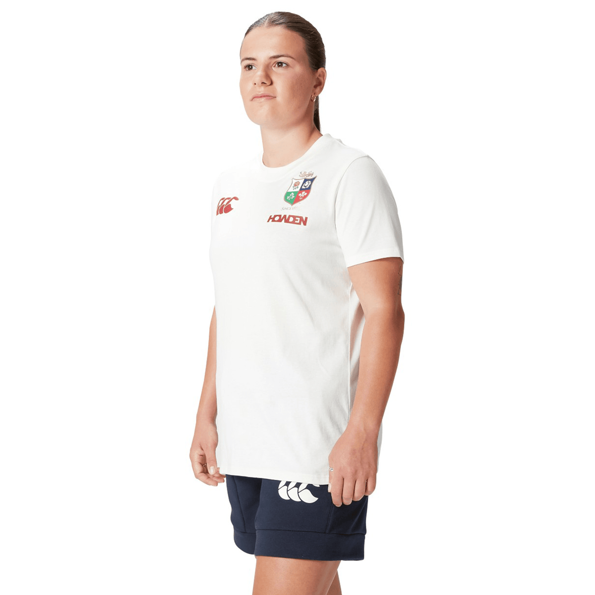 A dedicated rugby supporter stands confidently against a white background, wearing the Women's British &amp; Irish Lions 25 Cotton Jersey Tee by Canterbury, made of organic cotton with logos, and paired with classic black shorts.