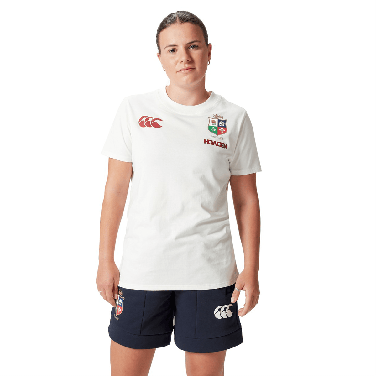 A rugby supporter proudly wearing the Women's British &amp; Irish Lions 25 Cotton Jersey Tee by Canterbury, featuring red British &amp; Irish Lions logos on a white background, and dark shorts stands against a white backdrop.