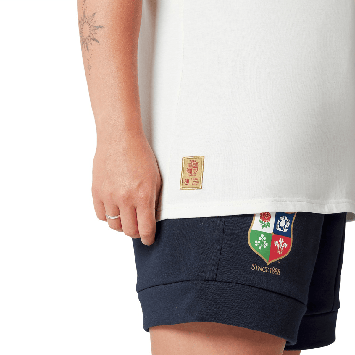 A rugby fan wearing the Women's British &amp; Irish Lions 25 Cotton Jersey Tee by Canterbury, paired with navy shorts adorned with vibrant embroidered patches, stands against a crisp white background. This outfit from Canterbury, crafted from organic cotton, effortlessly combines style and sustainability.