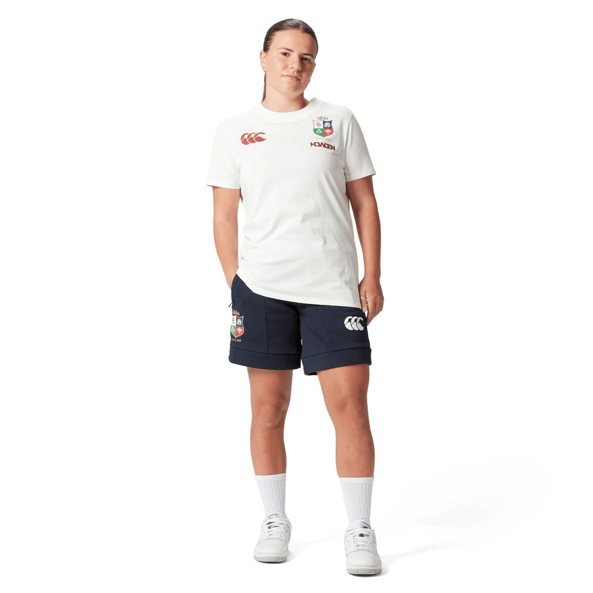 A rugby supporter wears a Women's British &amp; Irish Lions 25 Cotton Jersey Tee by Canterbury, paired with navy shorts featuring logos, complemented by white socks and sneakers against a plain white background. This outfit embodies the essence of the British &amp; Irish Lions.