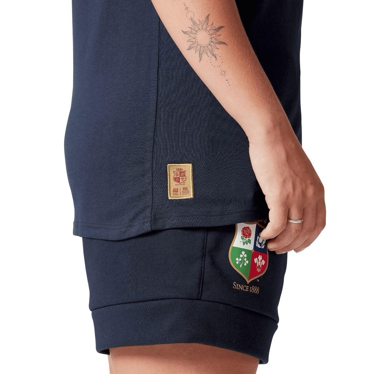 A close-up of a person wearing the Women's British &amp; Irish Lions 25 Cotton Jersey Tee by Canterbury in navy, featuring a sun tattoo on the arm and a patch with four emblems on the leg, captures the essence of every rugby supporter. Made from organic cotton, this jersey embodies sustainable style while celebrating the spirit of the British &amp; Irish Lions.