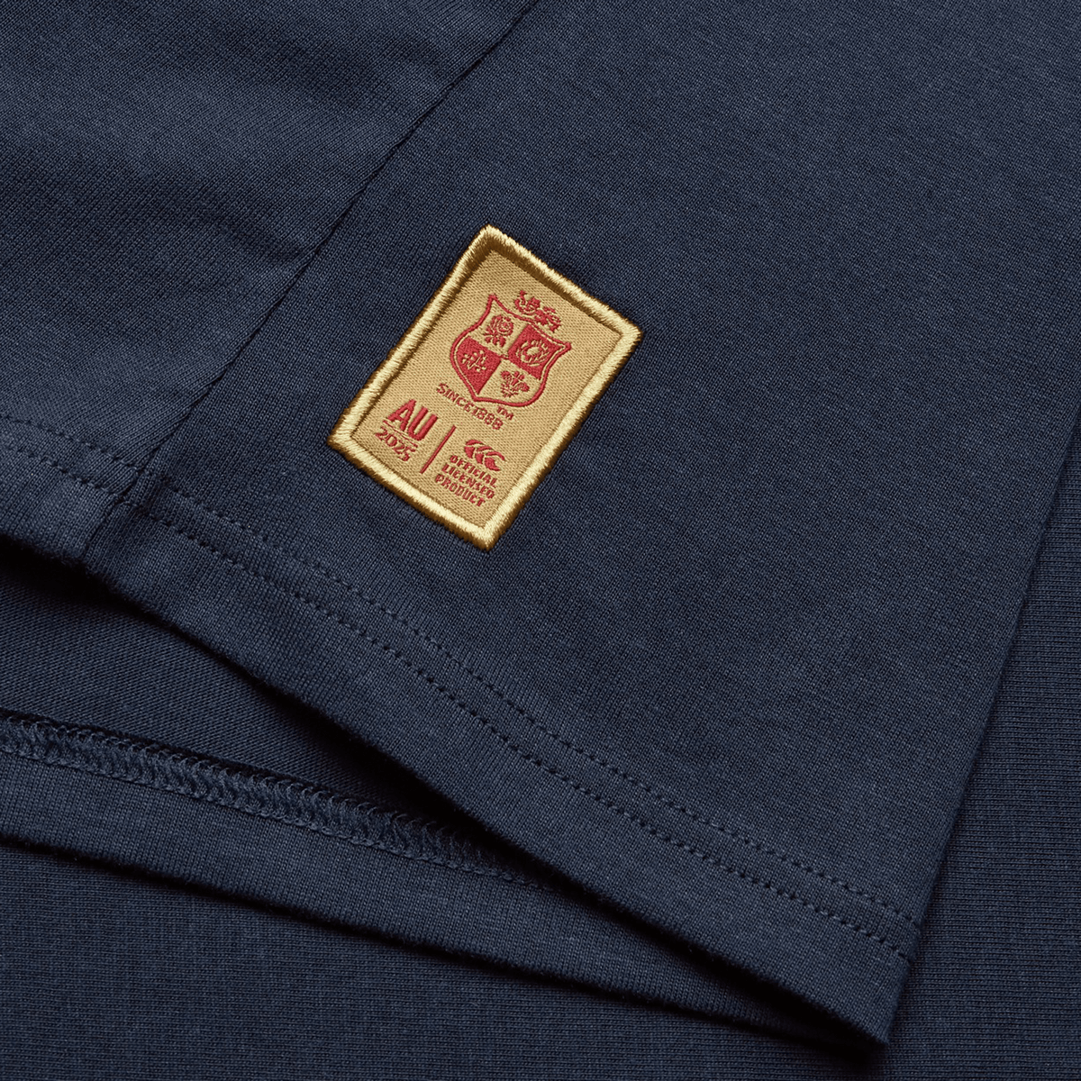A Women's British &amp; Irish Lions 25 Cotton Jersey Tee by Canterbury, in navy blue, is crafted from organic cotton and features a gold and red stitched label adorned with a crest, the text "AU 2025," along with additional small details underneath, making it perfect for any rugby supporter or fan of the British &amp; Irish Lions.