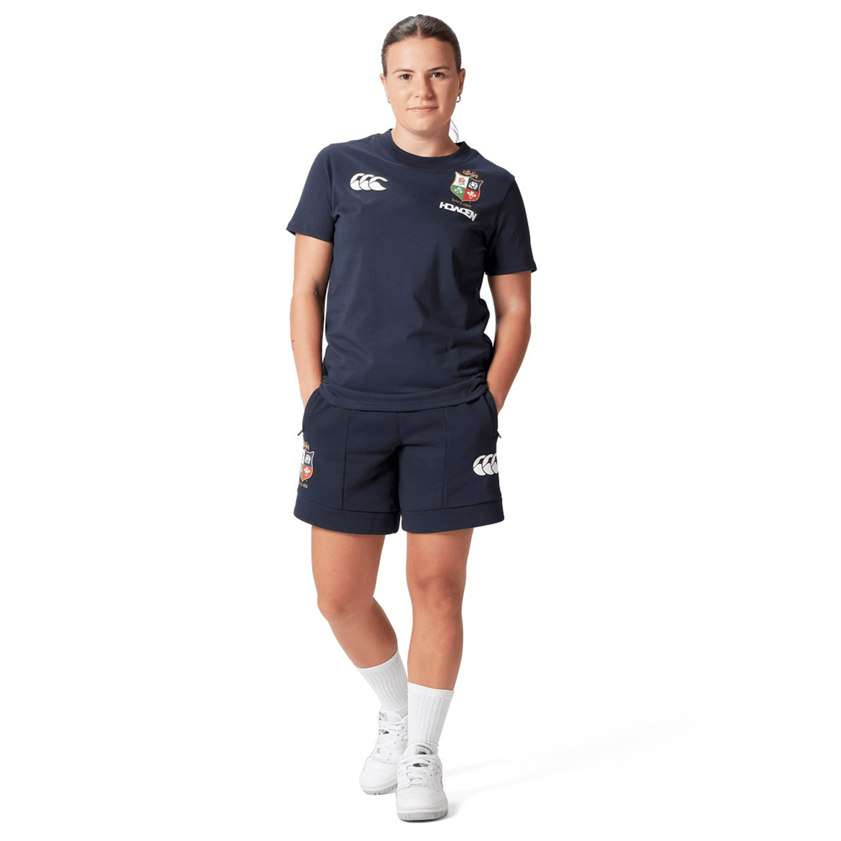 A rugby supporter stands confidently in the Women's British &amp; Irish Lions 25 Cotton Jersey Tee by Canterbury, adorned with logo patches. Complemented by white socks and shoes, this ensemble reflects their allegiance. The clean, white background highlights the sporty flair and dedication of a true British &amp; Irish Lions fan.