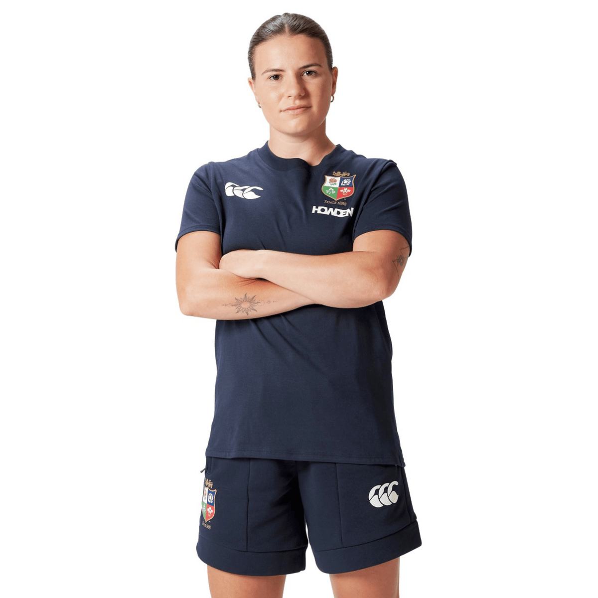 A rugby supporter in navy sportswear stands confidently with arms crossed, showcasing a Women's British &amp; Irish Lions 25 Cotton Jersey Tee by Canterbury and shorts featuring logos. This attire, made from organic cotton, proudly represents the spirit of the British &amp; Irish Lions.