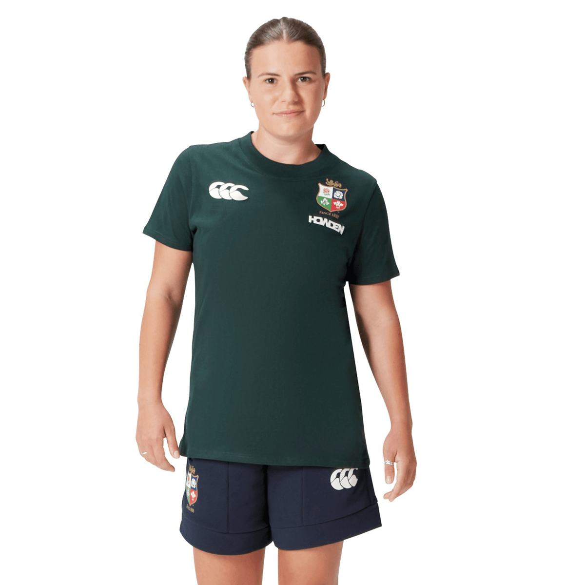 A rugby supporter wearing the Women's British &amp; Irish Lions 25 Cotton Jersey Tee by Canterbury, featuring a dark green color and made from organic cotton, paired with navy shorts adorned with logos, stands against a white background.