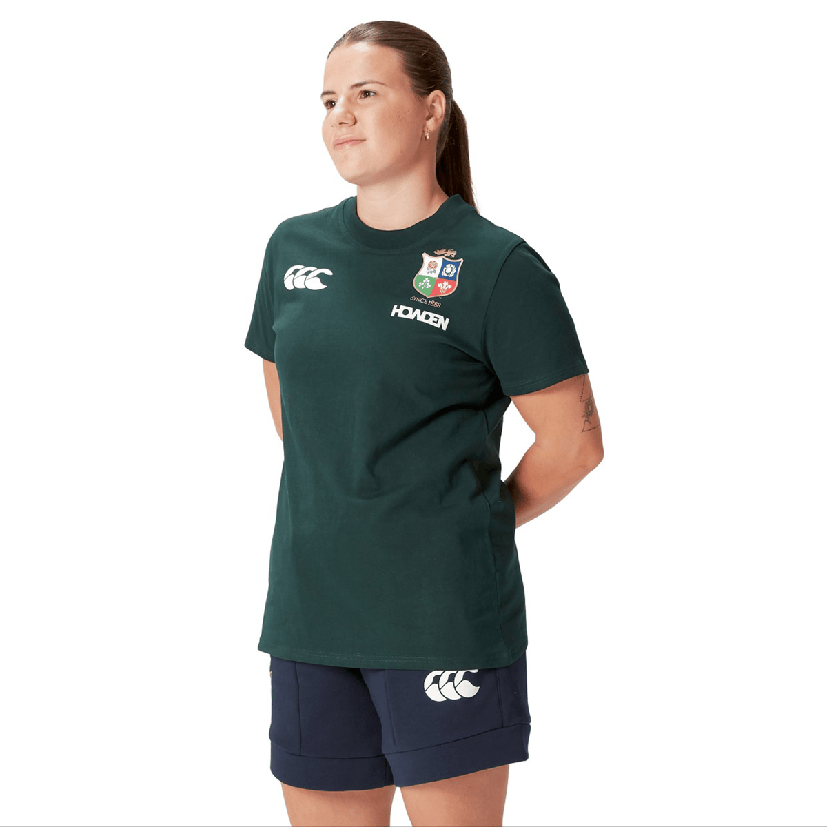 A rugby supporter proudly stands in a Women's British &amp; Irish Lions 25 Cotton Jersey Tee by Canterbury, with navy shorts, hands clasped behind their back, glancing to the side.