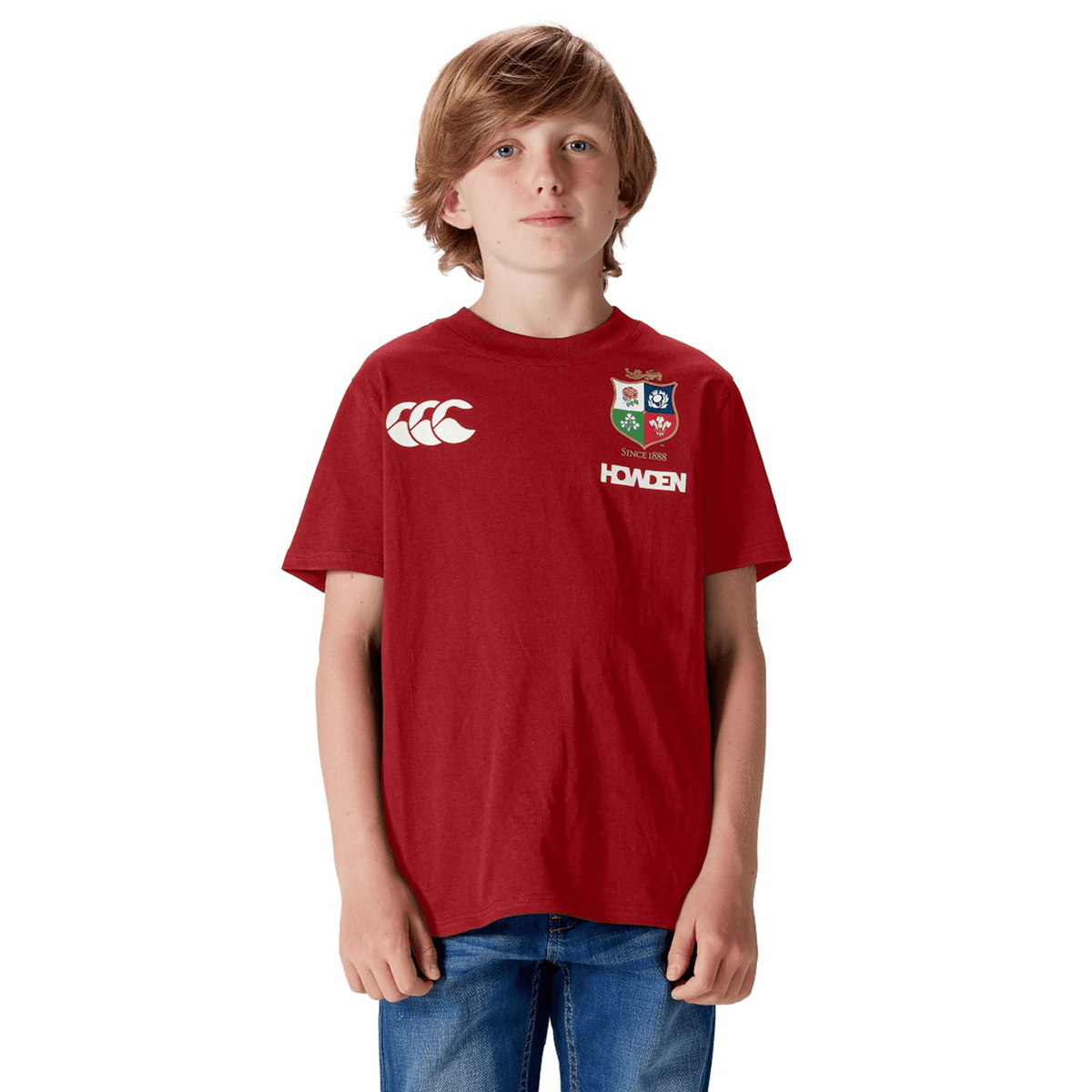 A rugby fan proudly poses against a white backdrop, wearing a Youth British &amp; Irish Lions 2025 Cotton Jersey Tee by Canterbury in red and pairing it with blue jeans.