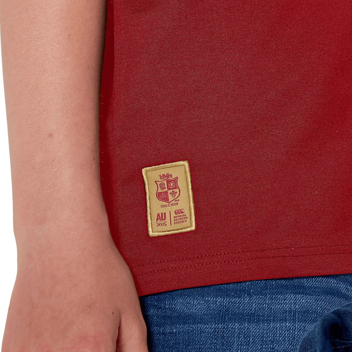A close-up shows a rugby fan in the Youth British &amp; Irish Lions 2025 Cotton Jersey Tee by Canterbury, made of organic cotton. It features a beige &quot;AU 2025&quot; logo patch. Paired with blue jeans, their hand casually rests near the shirt hem.