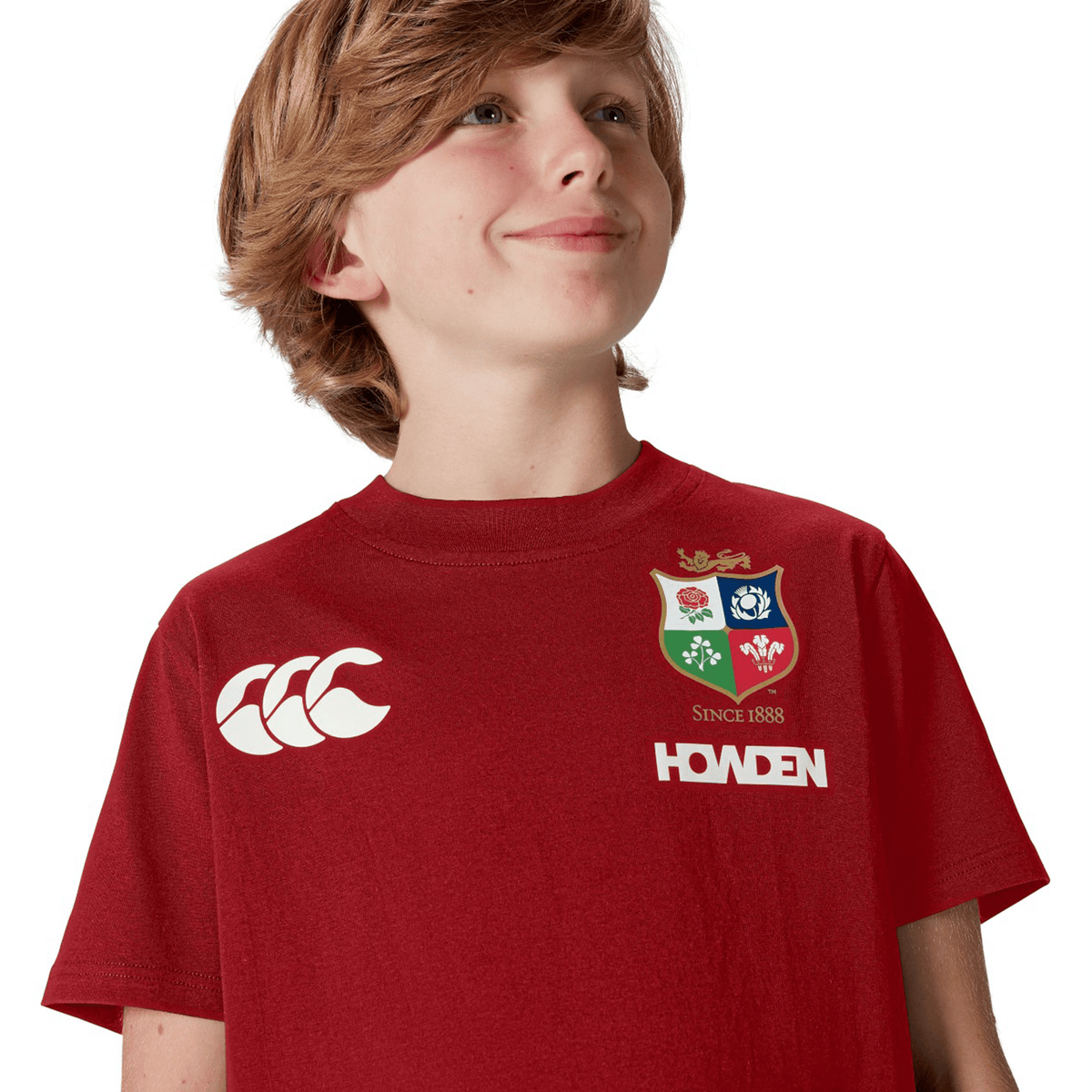 A young rugby fan, wearing a Canterbury Youth British &amp; Irish Lions 2025 Cotton Jersey Tee in red with a logo and text, gazes upwards with a slight smile, capturing the Lion&#39;s spirit.