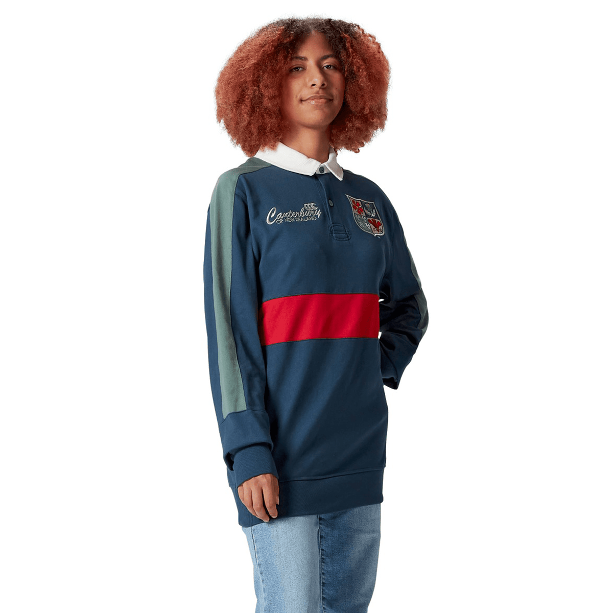 A person with curly hair poses confidently, wearing the British &amp; Irish Lions 2025 Heritage Jersey by Canterbury, featuring a navy design with a red stripe and emblem, embodying the classic Canterbury style, one hand tucked casually in their pocket.