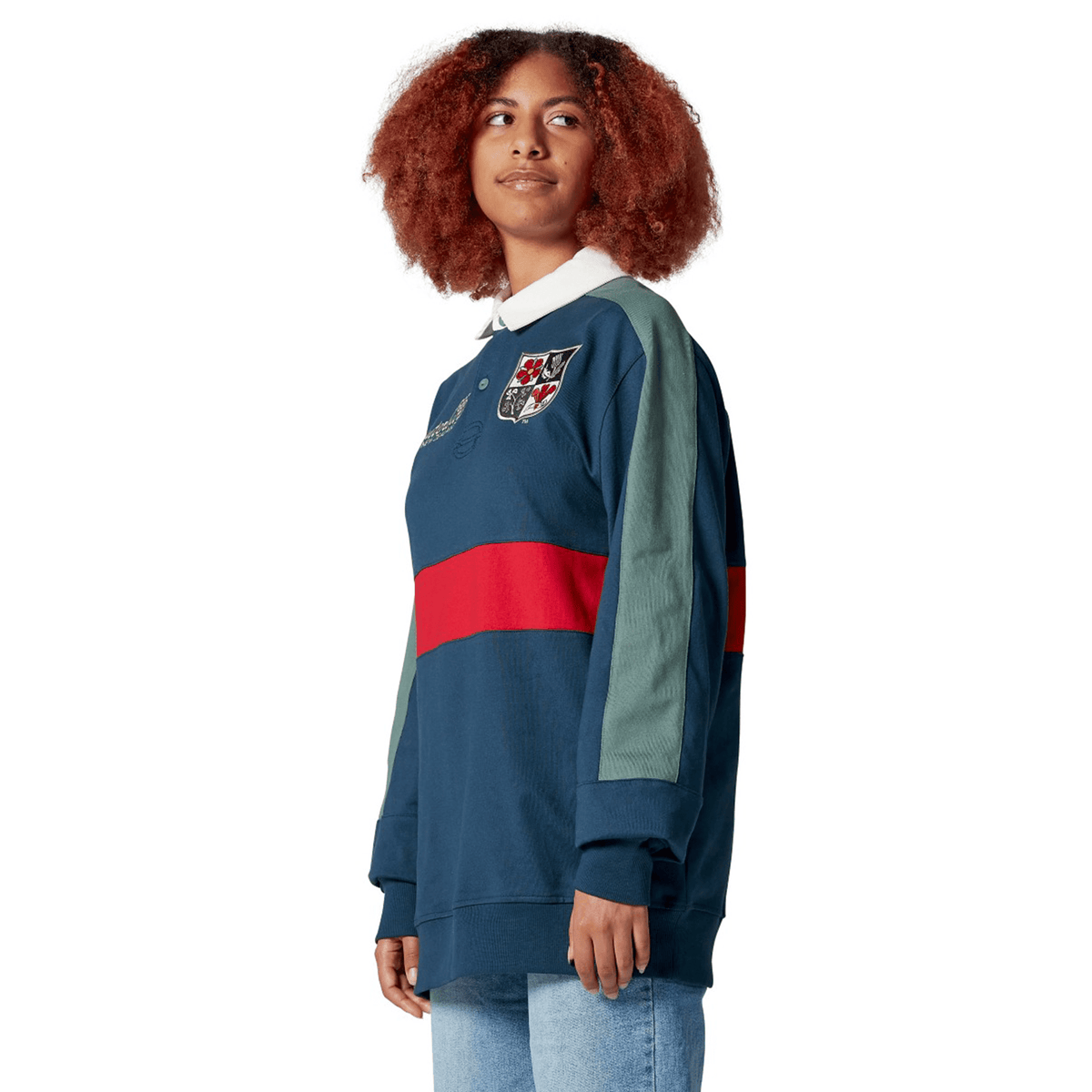 A woman with curly hair wears the British &amp; Irish Lions 2025 Heritage Jersey by Canterbury, showcasing a blue and green design with a red stripe and logo, paired with jeans. She gazes thoughtfully to the side.