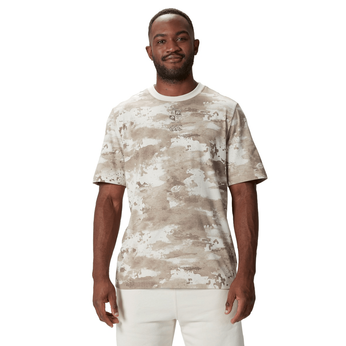 A man wears the British &amp; Irish Lions 2025 Camo Tee by Canterbury, styled with crisp white pants, standing against a pristine white background.