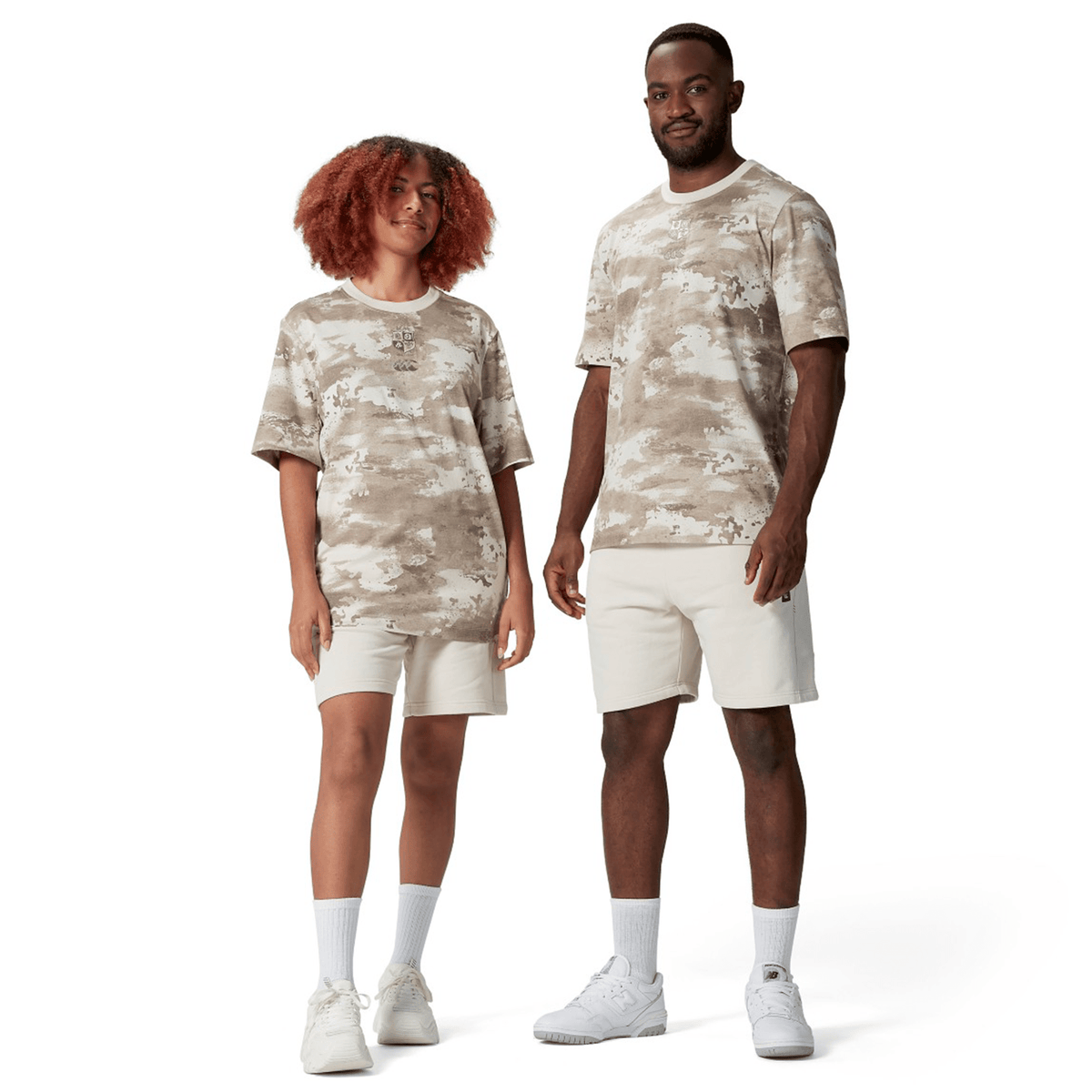 Two individuals stand on a plain white background, each wearing the British &amp; Irish Lions 2025 Camo Tee by Canterbury, paired with beige desert taupe shorts and crisp white sneakers.