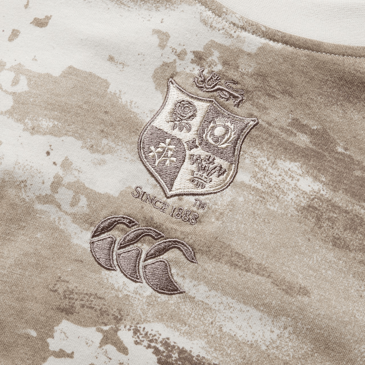 A close-up of a Desert Taupe camo tee, part of the British &amp; Irish Lions 2025 collection by Canterbury, showcases the British and Irish Lions logo, the text "Since 1888," and the Canterbury logo.