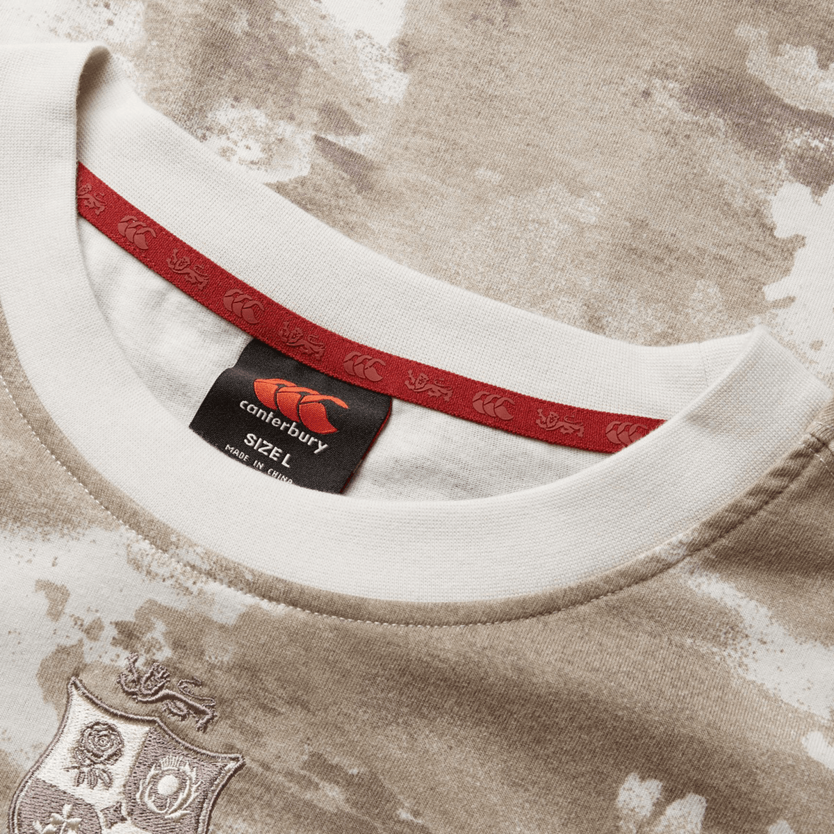A detailed view of the British &amp; Irish Lions 2025 Camo Tee by Canterbury shows a Desert Taupe and white pattern, complete with a red tag displaying the Canterbury logo and size "L." The fabric is adorned with an elegant crest design, evocative of the British &amp; Irish Lions.