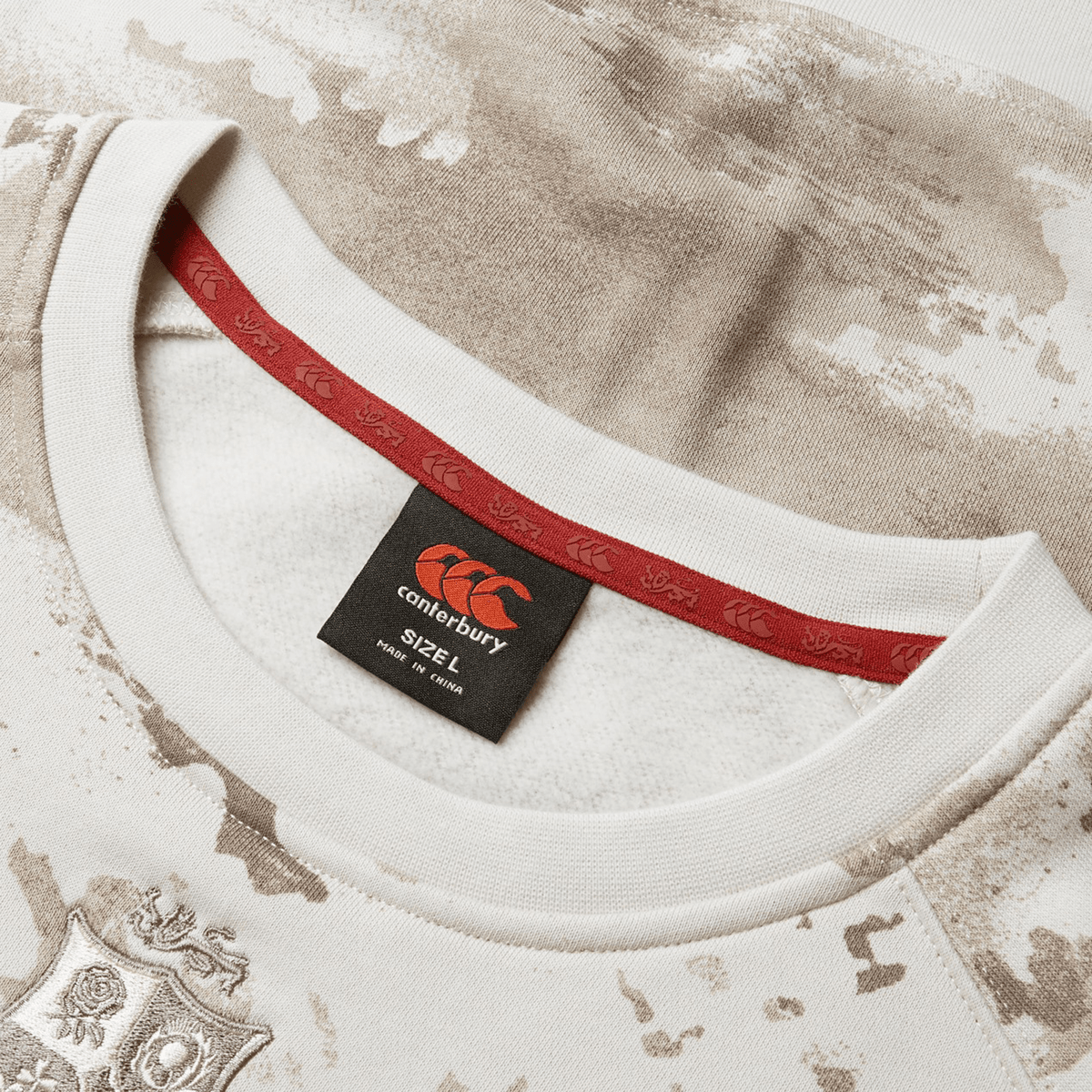 Close-up of a beige and white camouflage patterned sports jersey from Canterbury, reminiscent of the British &amp; Irish Lions 25 Camo Crew Sweatshirt. It includes a red inner collar with logo design and a visible "2XL" size label on the neck tag, reflecting the essence of British &amp; Irish Lions gear.