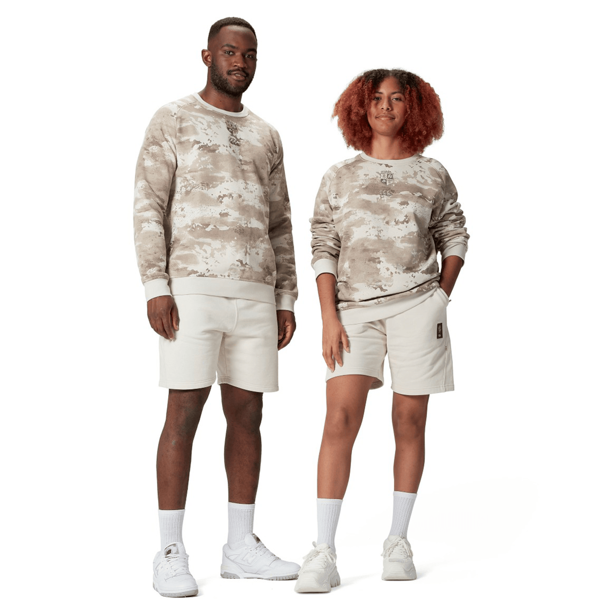 Two individuals pose side by side, donning identical beige British &amp; Irish Lions 25 Camo Crew Sweatshirts by Canterbury, paired with shorts. Both are wearing white sneakers and socks, embodying a relaxed and casual style. Their outfits reflect the stylish comfort associated with Canterbury, renowned for its British &amp; Irish Lions collections.