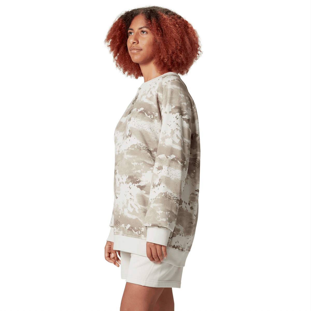 A person with curly hair confidently poses against a white background, modeling the British &amp; Irish Lions 25 Camo Crew Sweatshirt by Canterbury paired with light shorts.