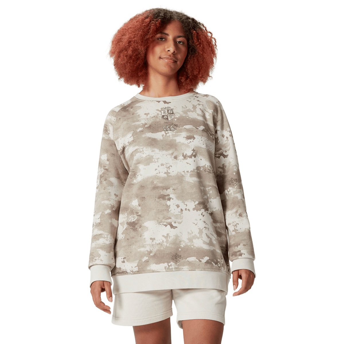 A person dressed in a beige British &amp; Irish Lions 25 Camo Crew Sweatshirt by Canterbury and shorts stands against a plain white background, subtly showcasing the style reminiscent of the brand.