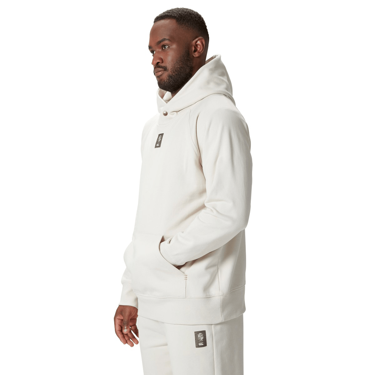 A man in a beige British &amp; Irish Lions 2025 Outback Fleece Hoodie by Canterbury and sweatpants stands with his hand in the pocket, exuding the effortless style characteristic of a Canterbury design.