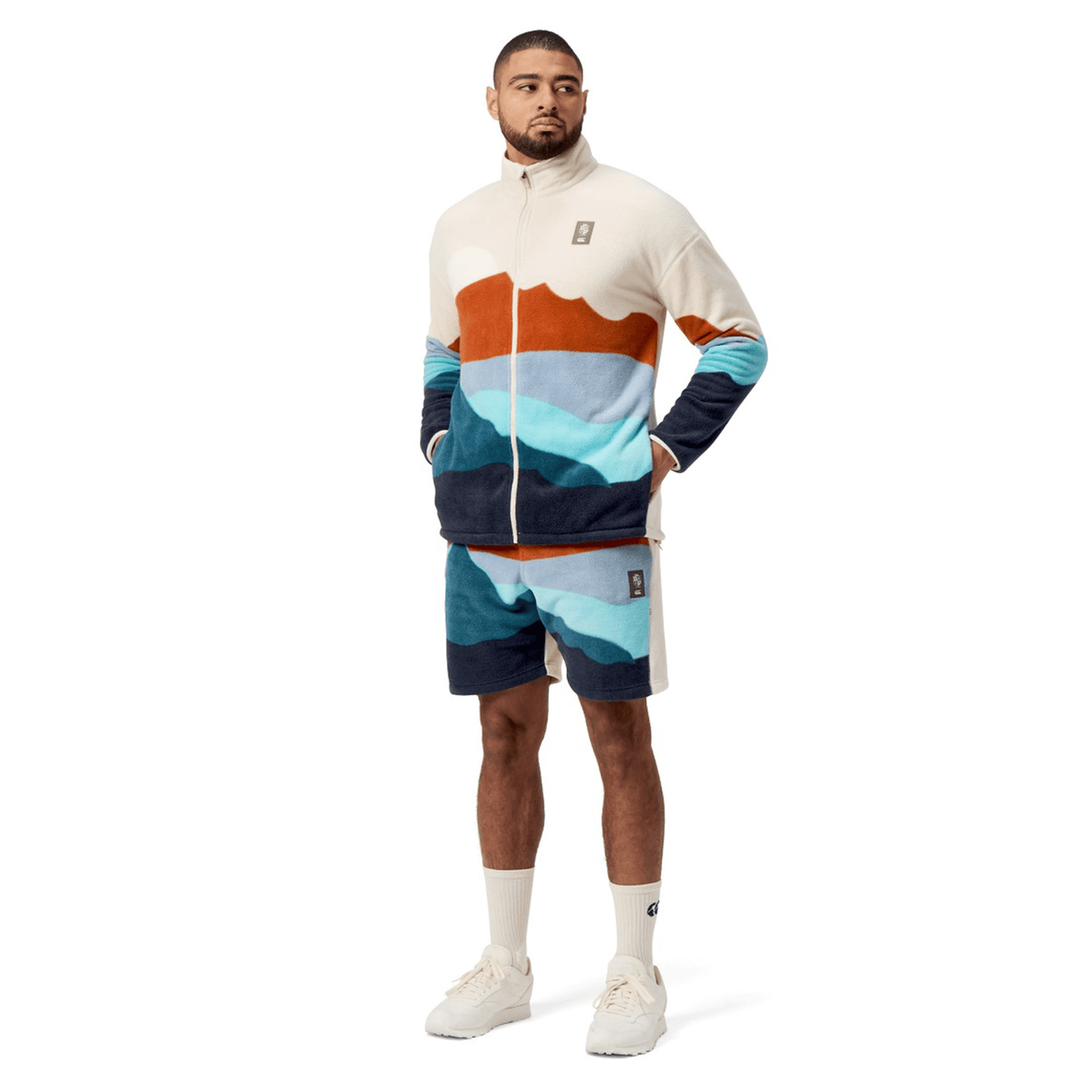 Dressed in a Canterbury British &amp; Irish Lions 2025 Outback Full-Zip Fleece Jacket, the man strikes a pose with hands tucked into his pockets, paired with shorts featuring a bold mountain pattern. He finishes off his outfit with white socks and sneakers, capturing an adventurous spirit that echoes the essence of the British &amp; Irish Lions.
