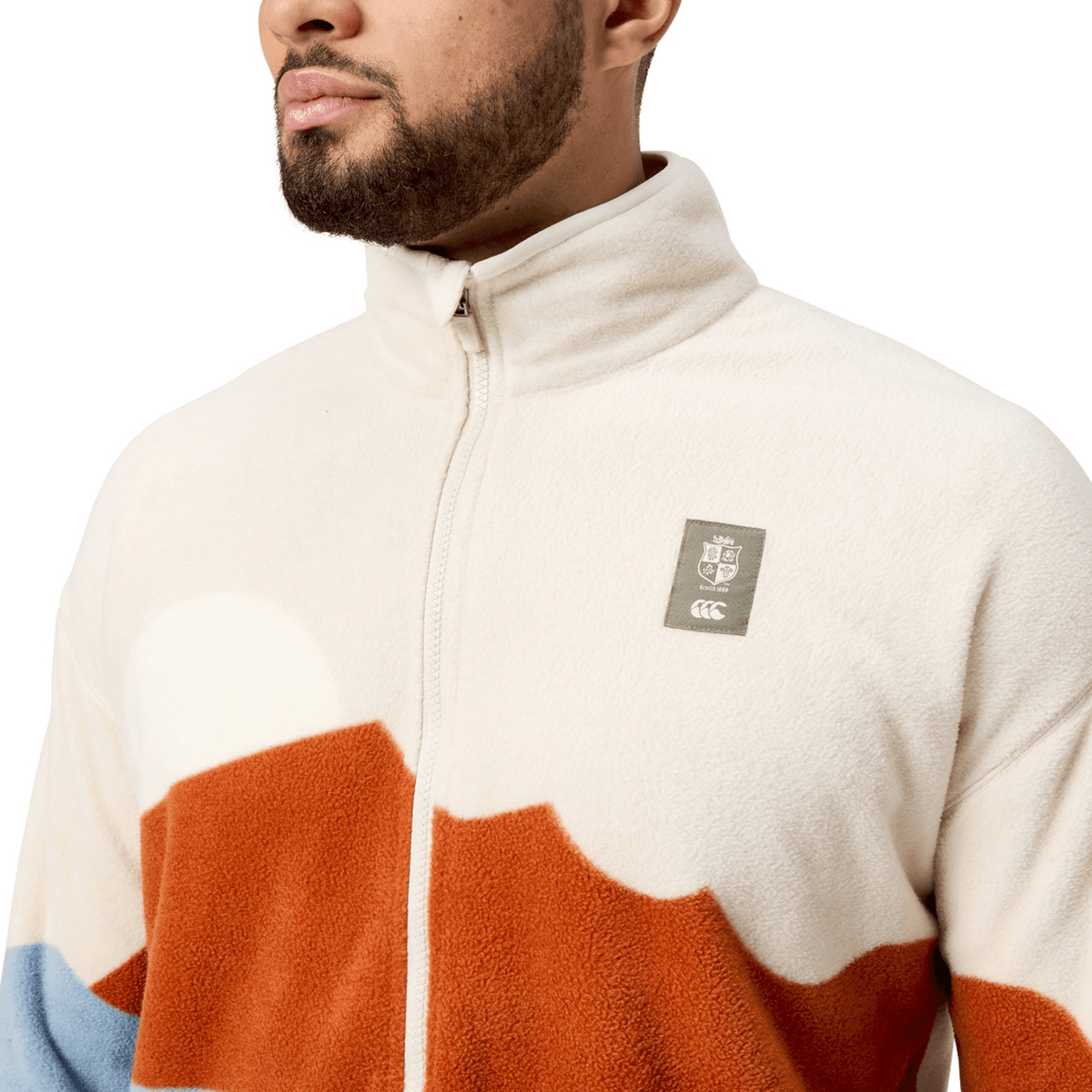 Someone is wearing a cream and rust-colored British &amp; Irish Lions 2025 Outback Full-Zip Fleece Jacket by Canterbury, featuring a high collar and prominently displaying the Canterbury crest logo on the chest.